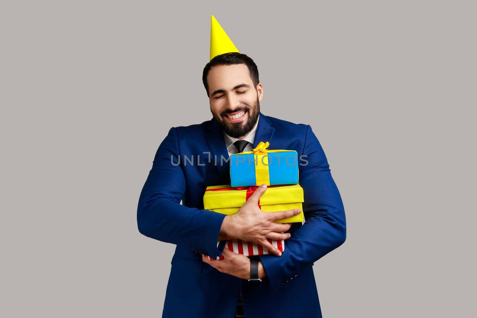 Extremely happy bearded man posing in party cone, holding embracing stack of presents, happy birthday, wearing official style suit. Indoor studio shot isolated on gray background.