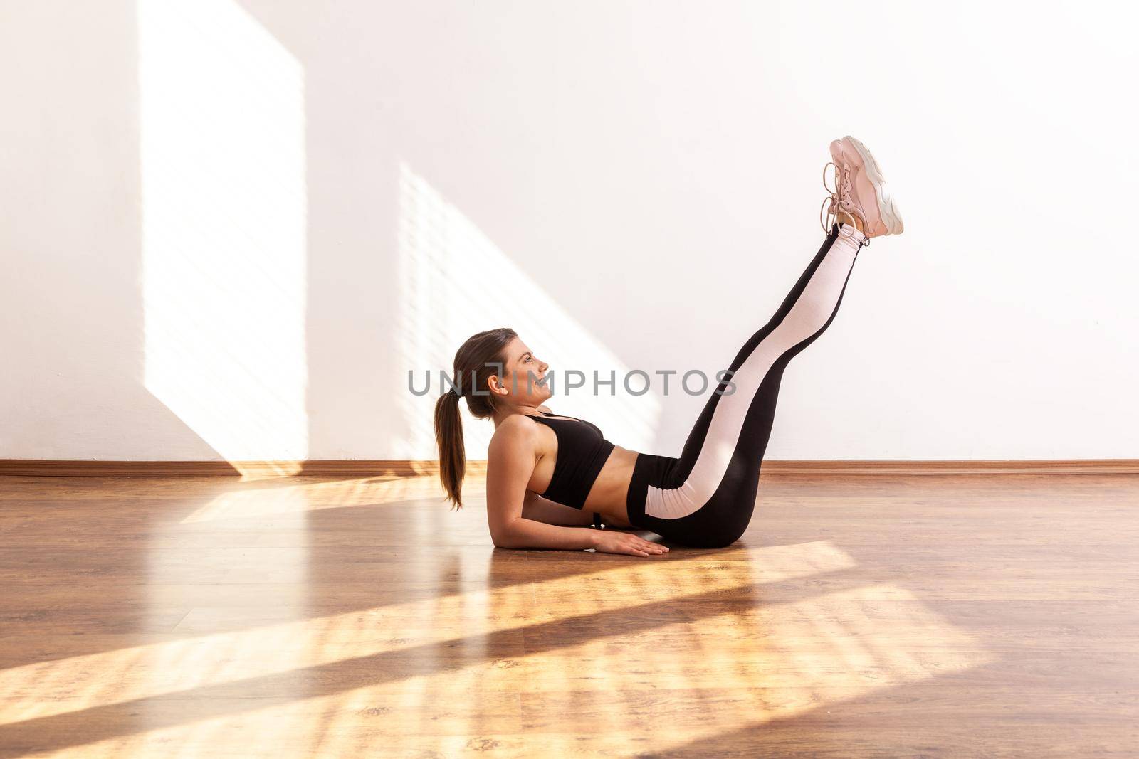 Assertive woman lying on floor raising legs, doing crunches, pumping up abdominal and spine muscles, wearing black sports top and tights. Full length studio shot illuminated by sunlight from window.