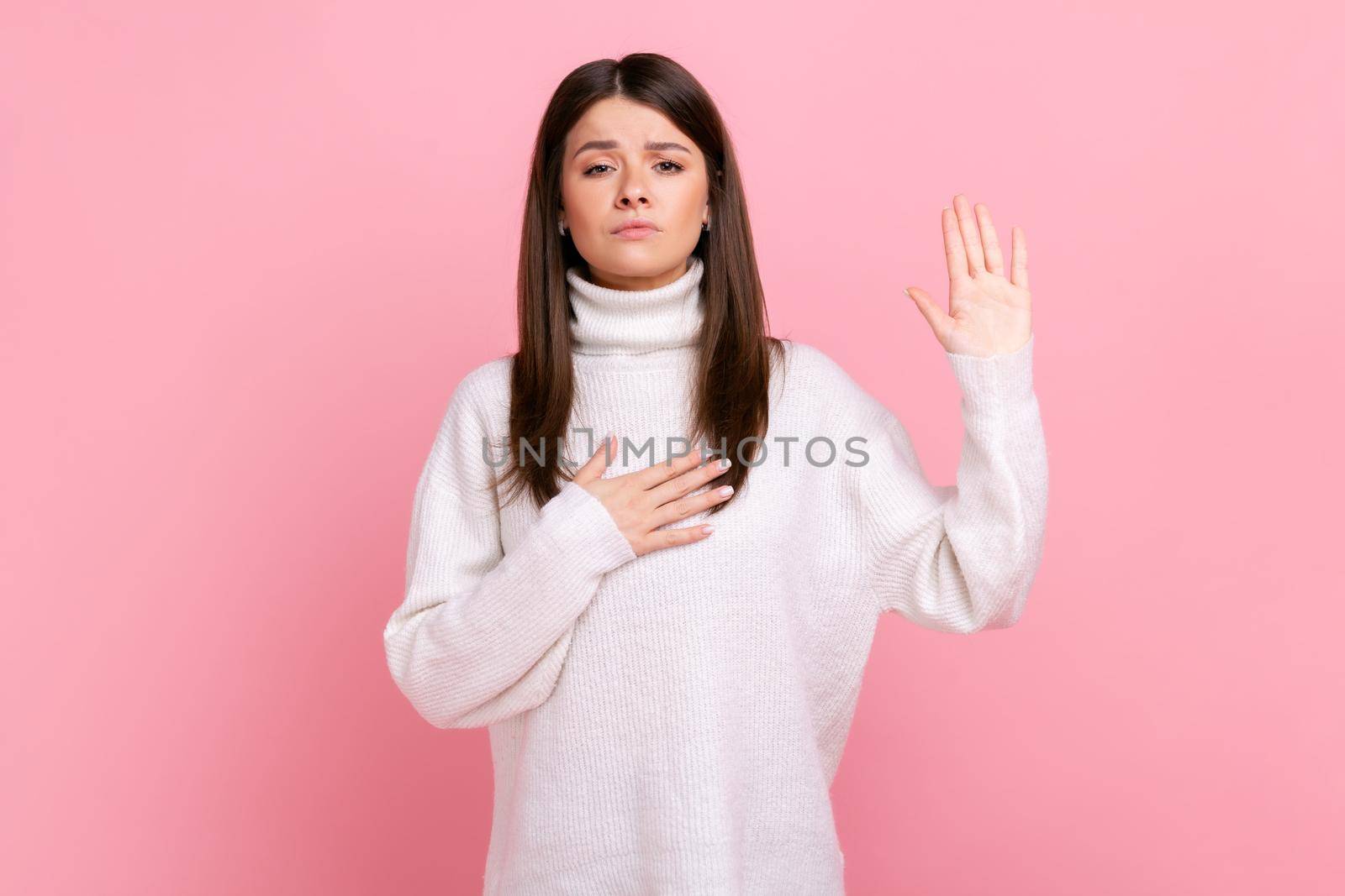 Portrait of honest young woman standing with hand on chest and fingers up, making loyalty promise, wearing white casual style sweater. Indoor studio shot isolated on pink background.