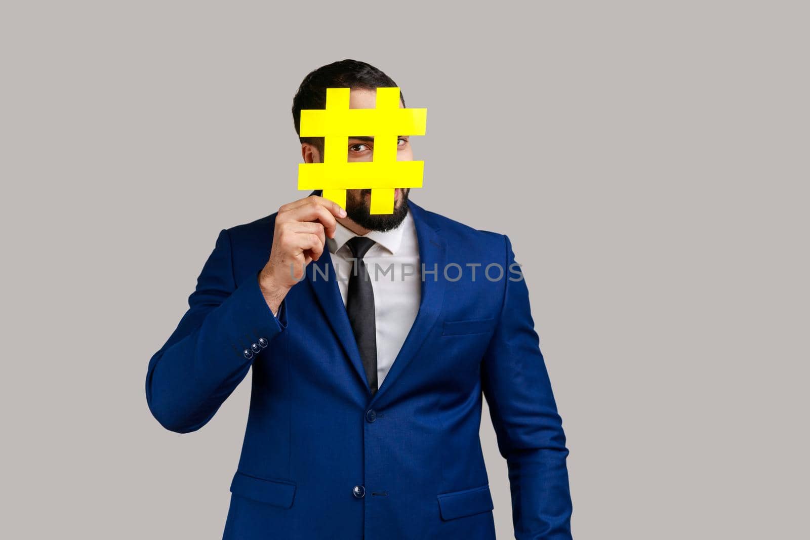Man covering face with social media hashtag symbol, recommending to follow trendy content, popular business blog, wearing official style suit. Indoor studio shot isolated on gray background.