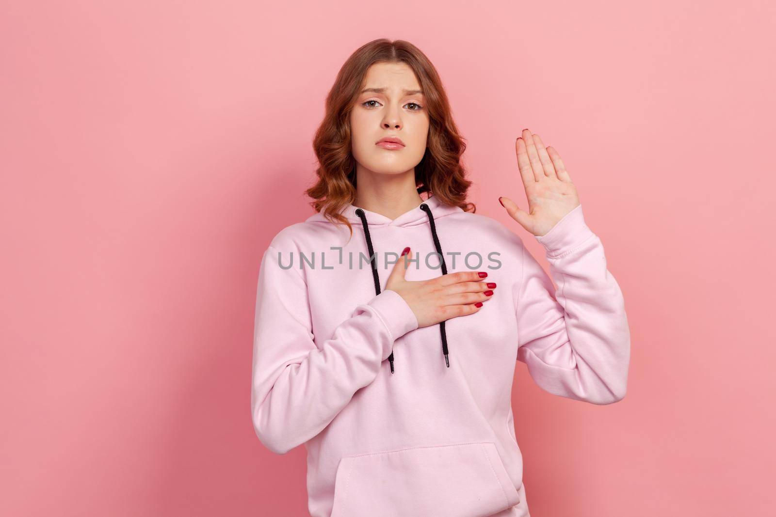 Portrait of sincere honest teen girl in hoodie holding hand up and on chest, pledging allegiance, taking oath with responsible expression by Khosro1