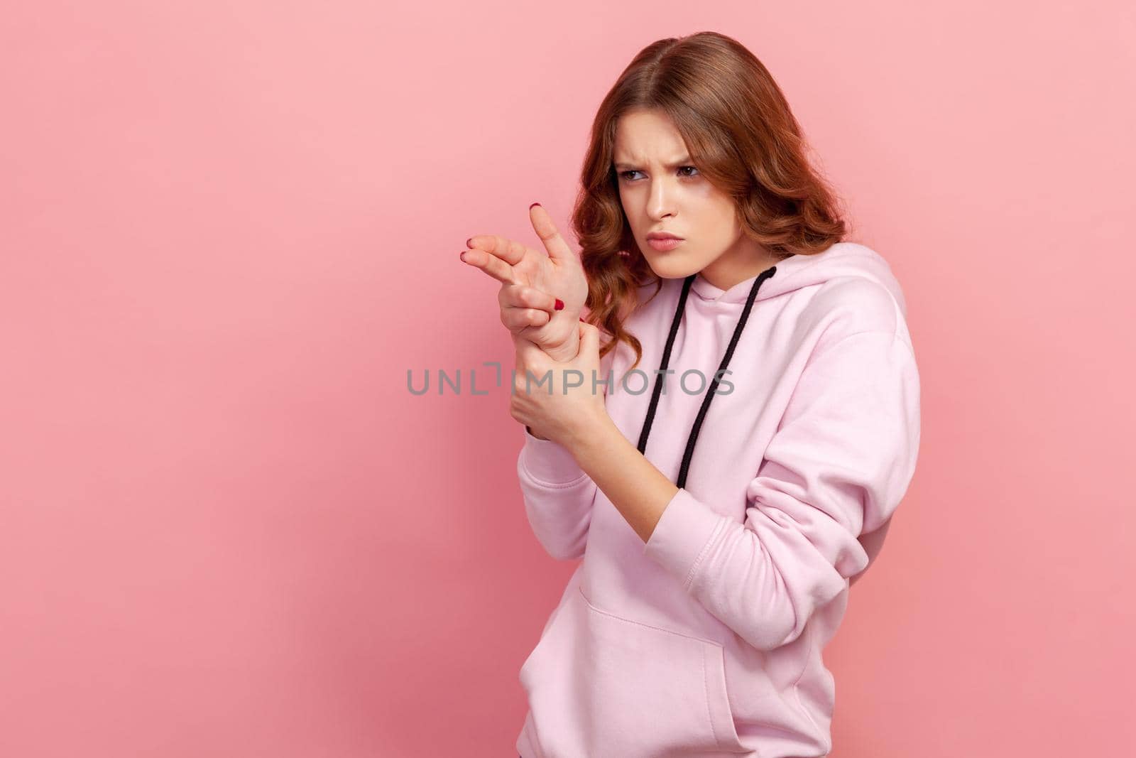 Portrait of curly-haired teen girl in hoodie pointing finger gun to camera, aiming and threatening to shoot with pistol hand gesture. Indoor studio shot isolated on pink background
