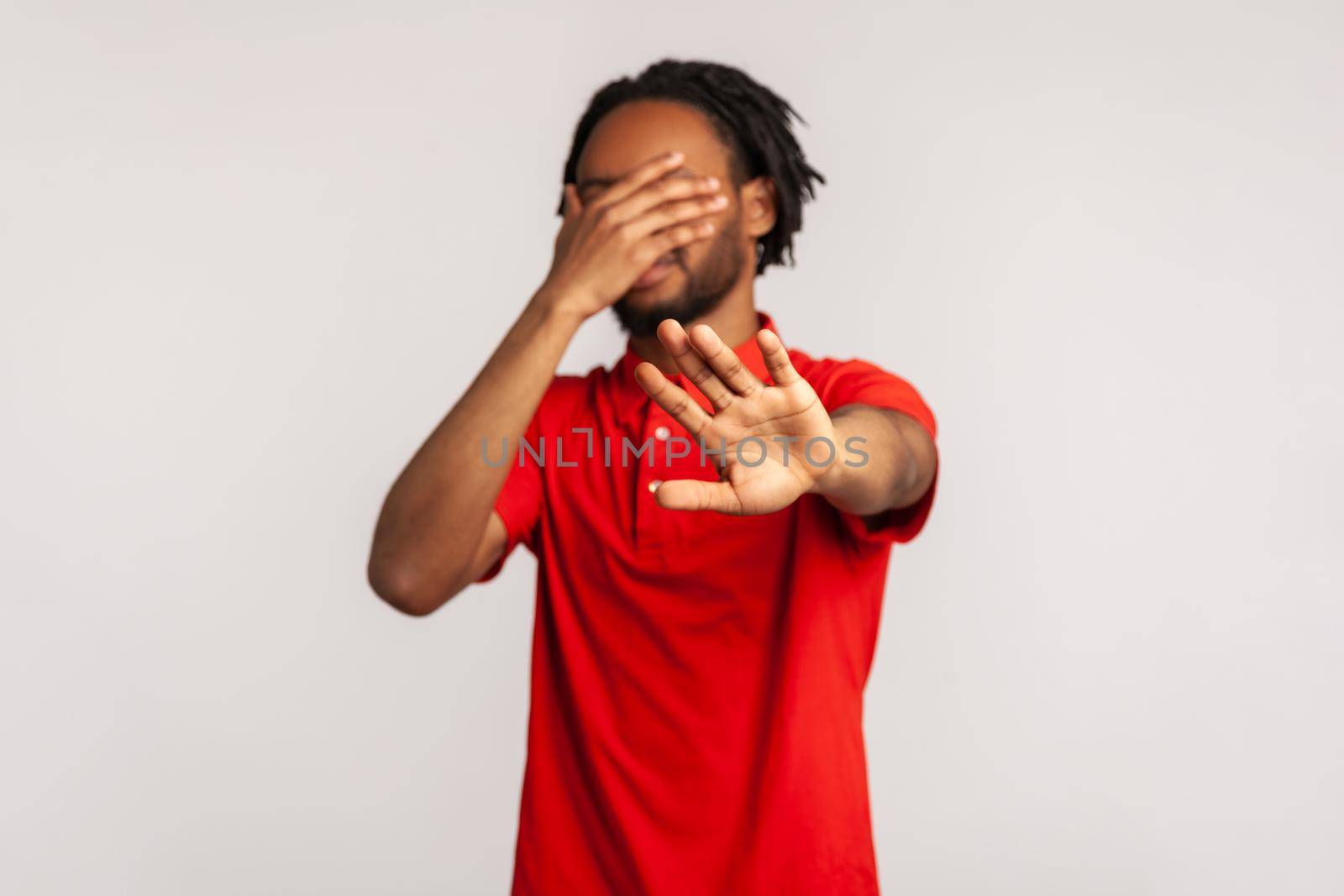 Stop, I don't want to see at this. Young bearded man with dreadlocks wearing red casual style T-shirt, covering his face and showing stop hand gesture. Indoor studio shot isolated on gray background.