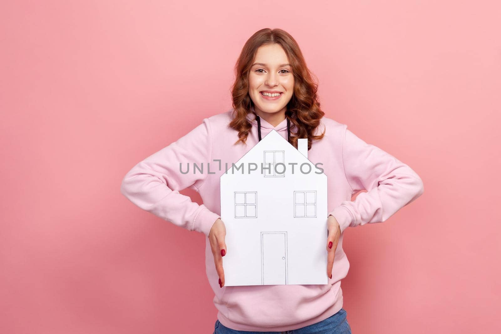 Portrait of excited curly haired teenage girl in hoodie holding paper house looking at camera with toothy smile, dreaming of own home. Indoor studio shot isolated on pink background