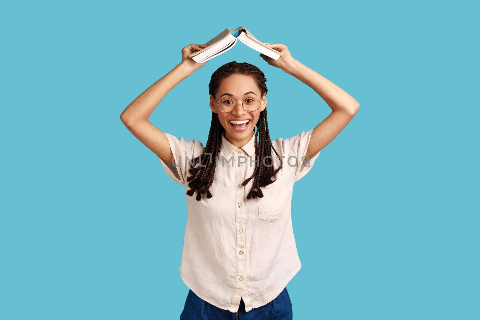 Portrait of positive woman with black dreadlocks and eyeglasses, standing holding book over head, studying and education, wearing white shirt. Indoor studio shot isolated on blue background.