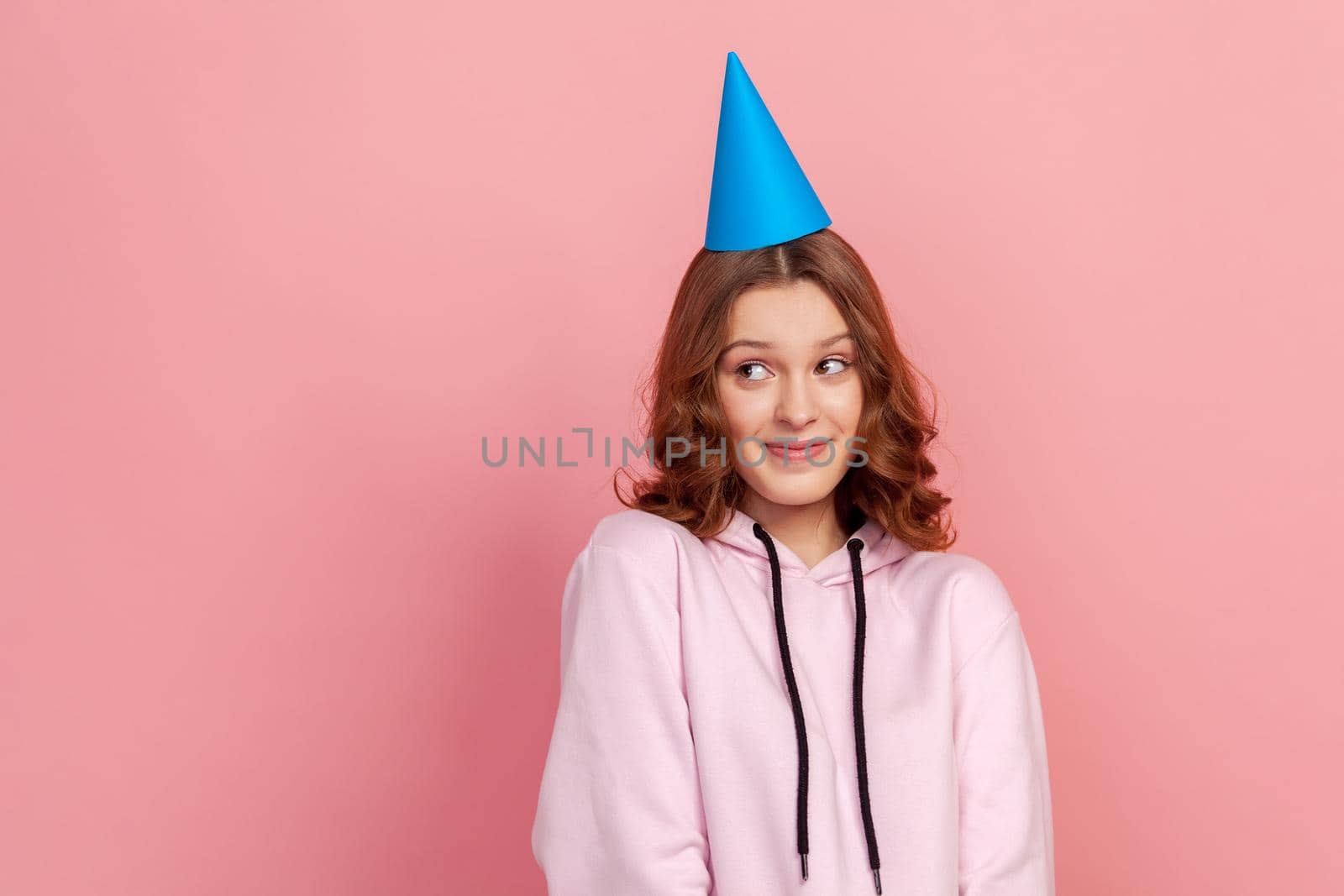 Portrait of shy curly haired teenage girl in hoodie and blue party hat looking up, birthday greetings. Indoor studio shot isolated on pink background