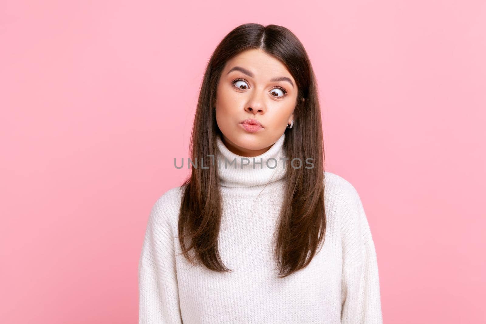 Idiot woman making silly humorous face with eyes crossed, showing comical silly brainless expression, wearing white casual style sweater. Indoor studio shot isolated on pink background.