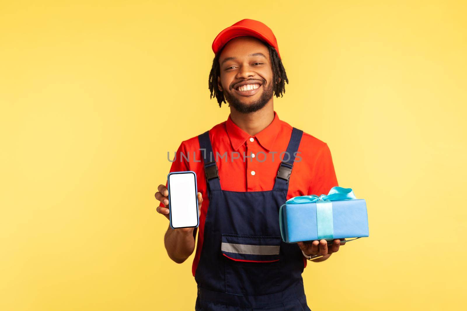 Portrait of happy smiling deliveryman in blue overalls, red T-shirt and cap holding present box and smart phone with blank screen for advertisement. Indoor studio shot isolated on yellow background.