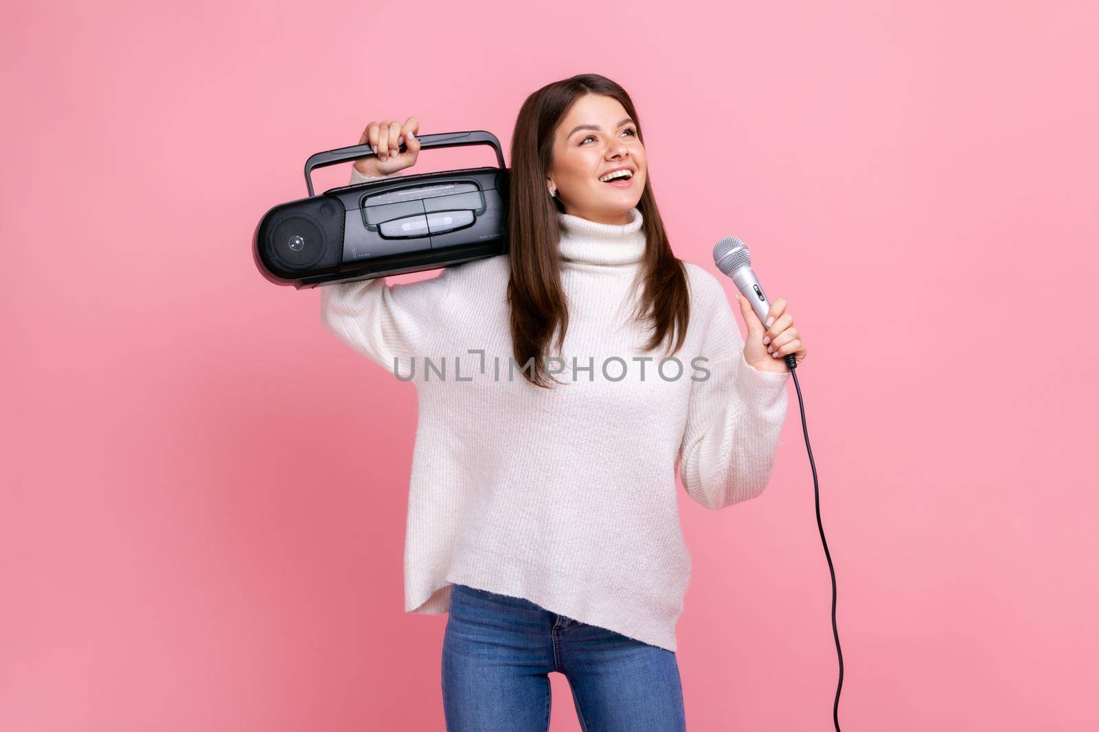 Portrait of happy positive female having fun, holding record player and singing via microphone, wearing white casual style sweater and jeans. Indoor studio shot isolated on pink background.