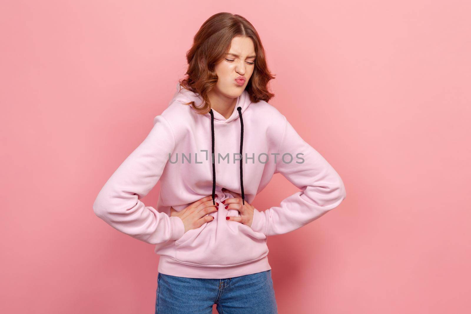 Portrait of upset teenager female in hoodie grimacing and holding belly, suffering abdominal pain and spasm, gastritis. Indoor studio shot isolated on pink background