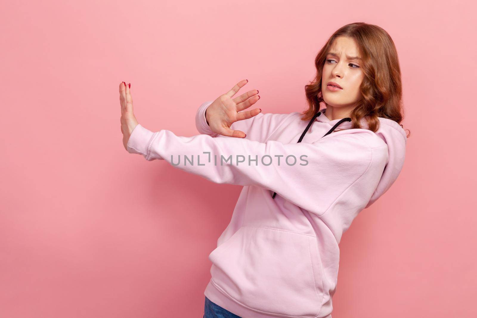 Side view of scared young girl standing with raised hands, gesturing stop, panicking looking frightened of danger, phobia. Indoor studio shot isolated on pink background