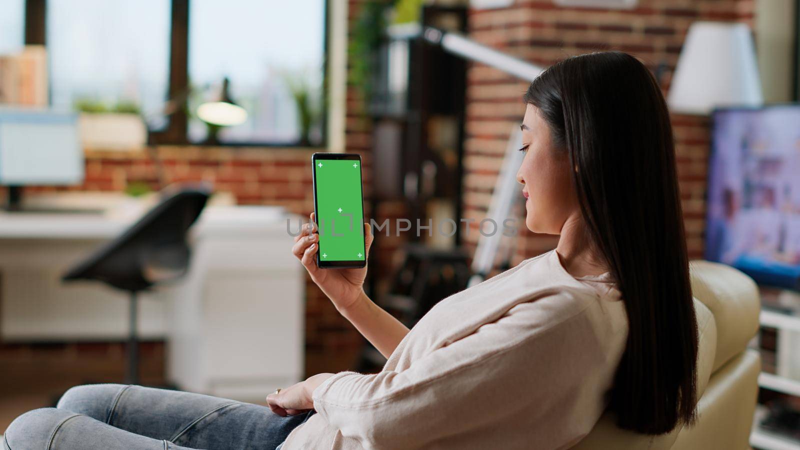 Young adult person sitting on couch with mobile cellphone having mockup chroma key background. Asian woman having smartphone with green screen display while sitting at home on sofa. Tripod shot
