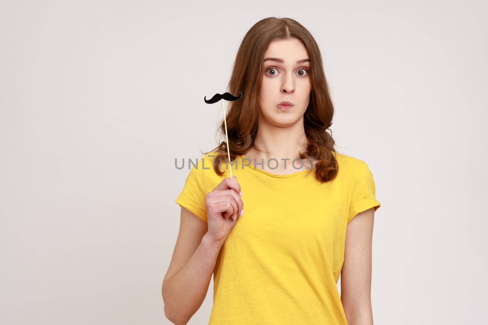 Portrait of astonished young female in yellow T-shirt holding black paper party mustache on stick, looking at camera with confused expression. Indoor studio shot isolated on gray background.