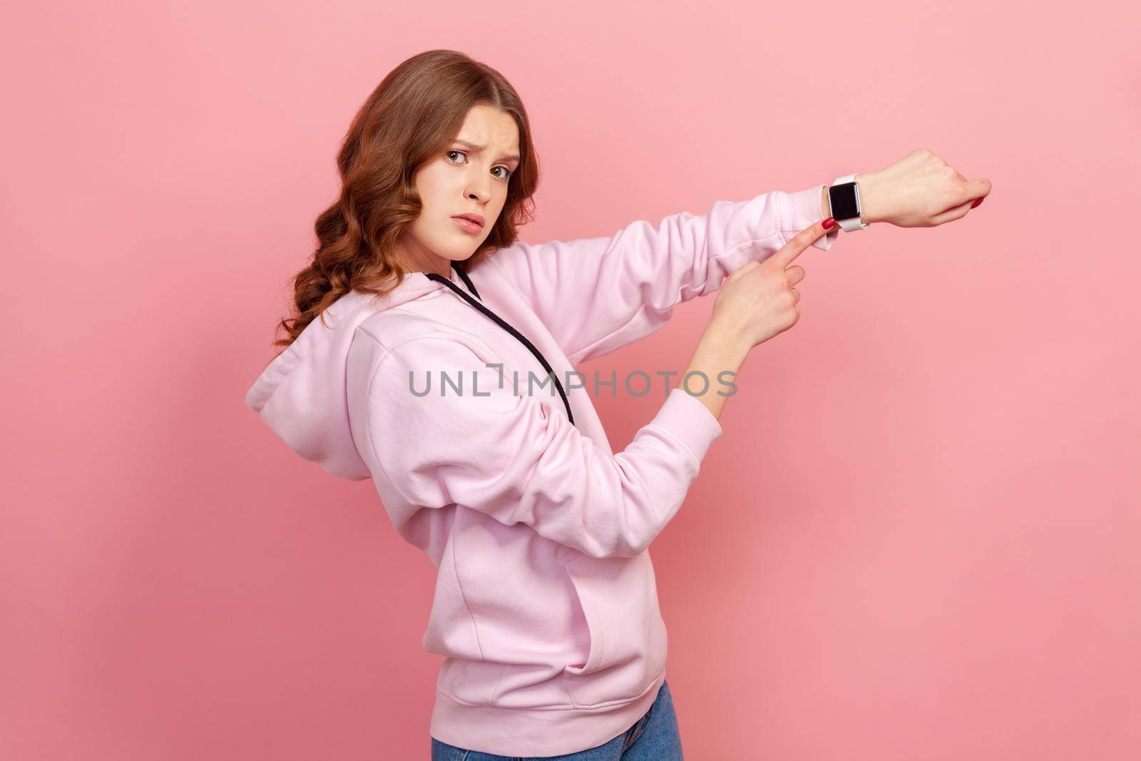 Portrait of displeased young brunette female in hoodie looking impatient and pointing wristwatch, warning of deadline, late hour. Indoor studio shot isolated on pink background