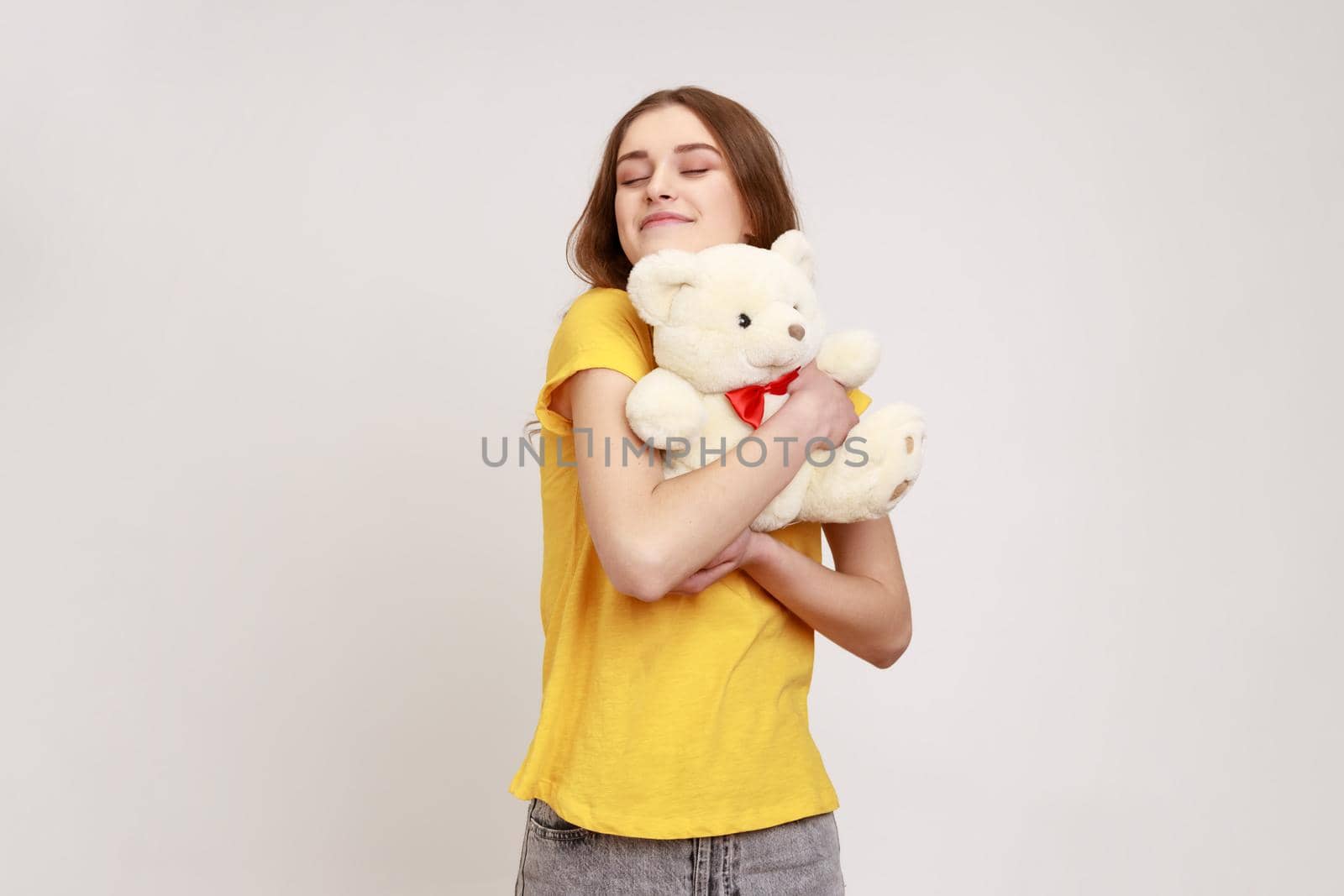 Satisfied happy teenager girl with brown hair in yellow casual T-shirt hugging cute white toy bear, closing eyes and dreaming, enjoying romantic gift. Indoor studio shot isolated on gray background.