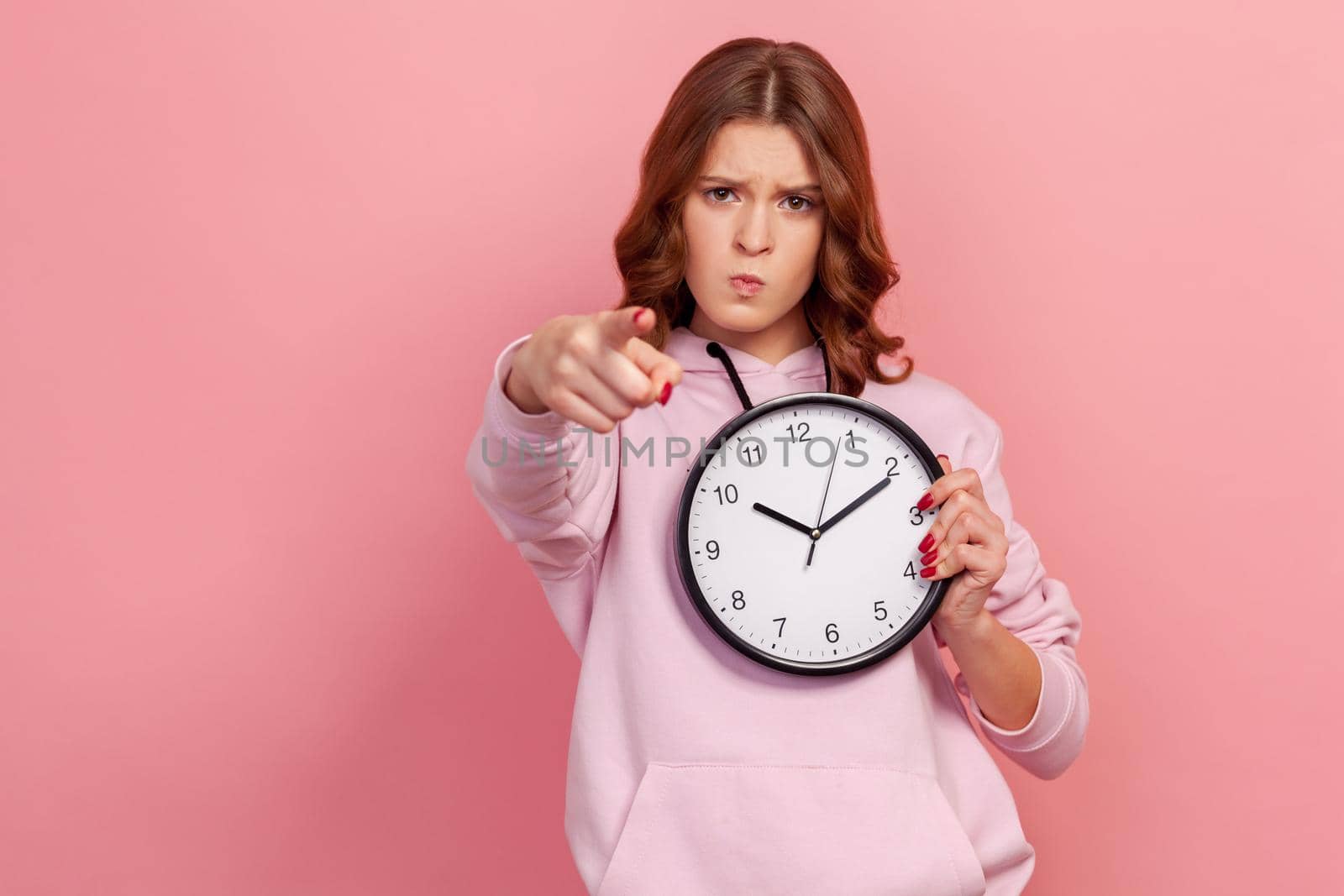 Portrait of angry serious teen girl in hoodie pointing finger at camera holding big wall clock in hand, time to action. Indoor studio shot isolated on pink background