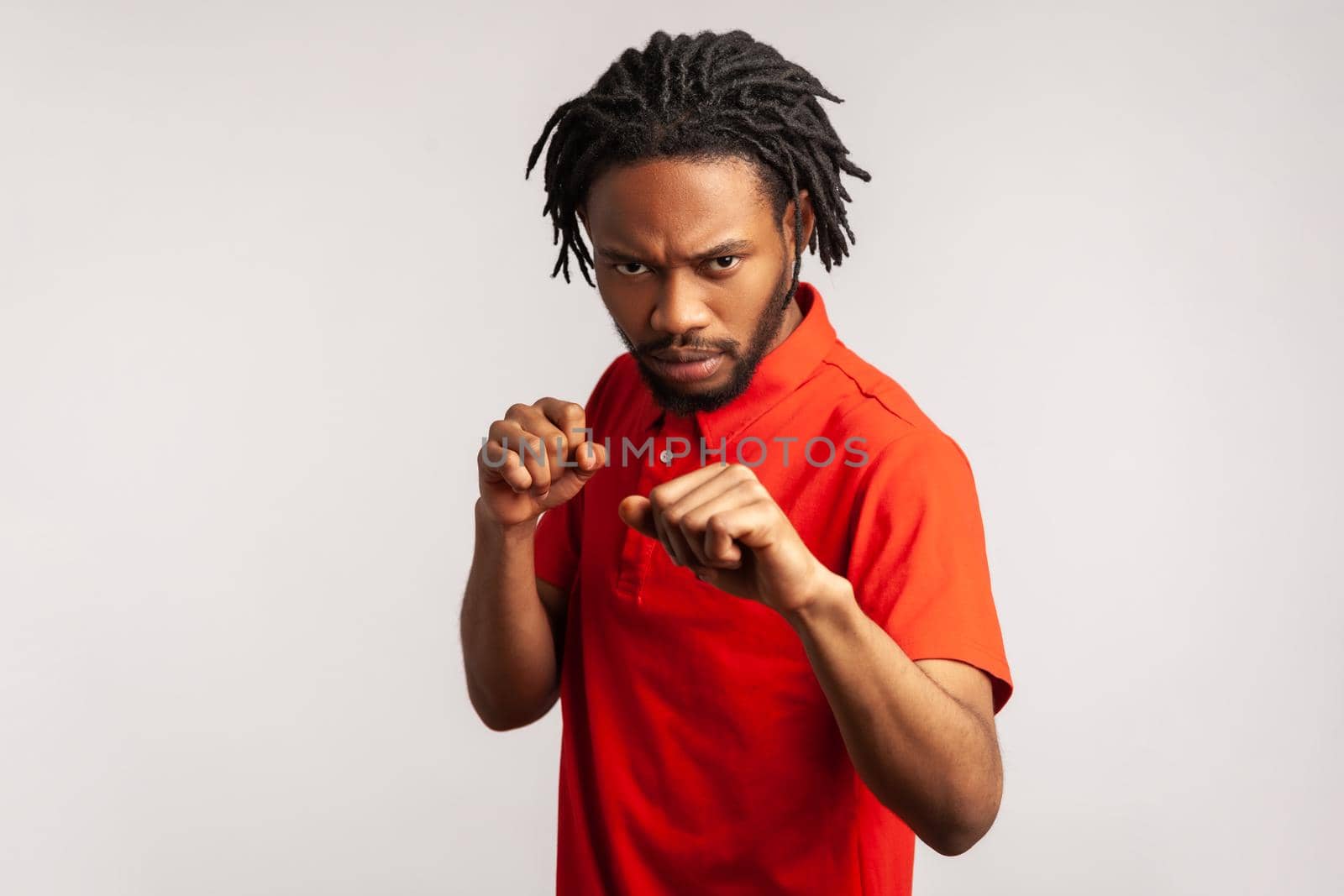 Aggressive bearded man with dreadlocks wearing red casual style T-shirt, holding clenched fists up ready to boxing, martial art trainer, self defense. Indoor studio shot isolated on gray background.