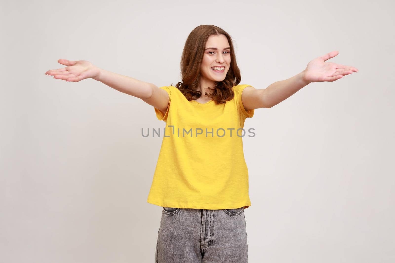 Come into my arms, free hugs. Portrait of adorable hospitable teenager girl yellow casual T-shirt smiling and reaching out hands, going to embrace. Indoor studio shot isolated on gray background.