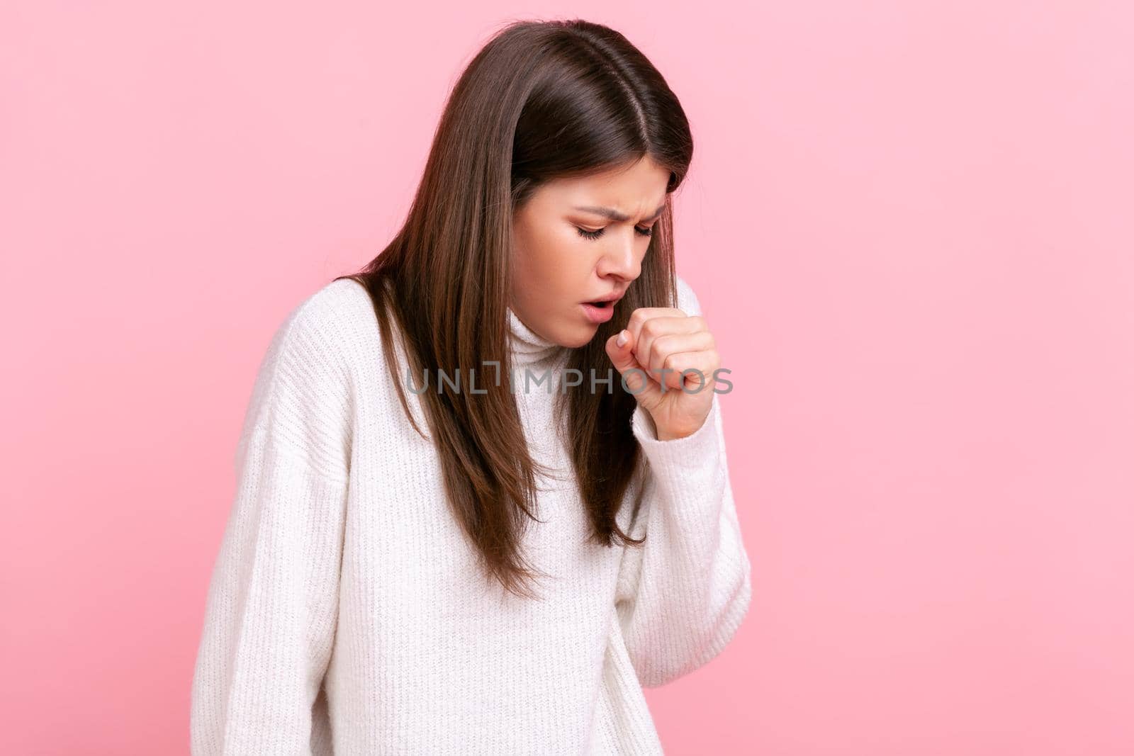 Profile portrait of young adult brunette sick woman coughing, catches cold, having high temperature, wearing white casual style sweater. Indoor studio shot isolated on pink background.