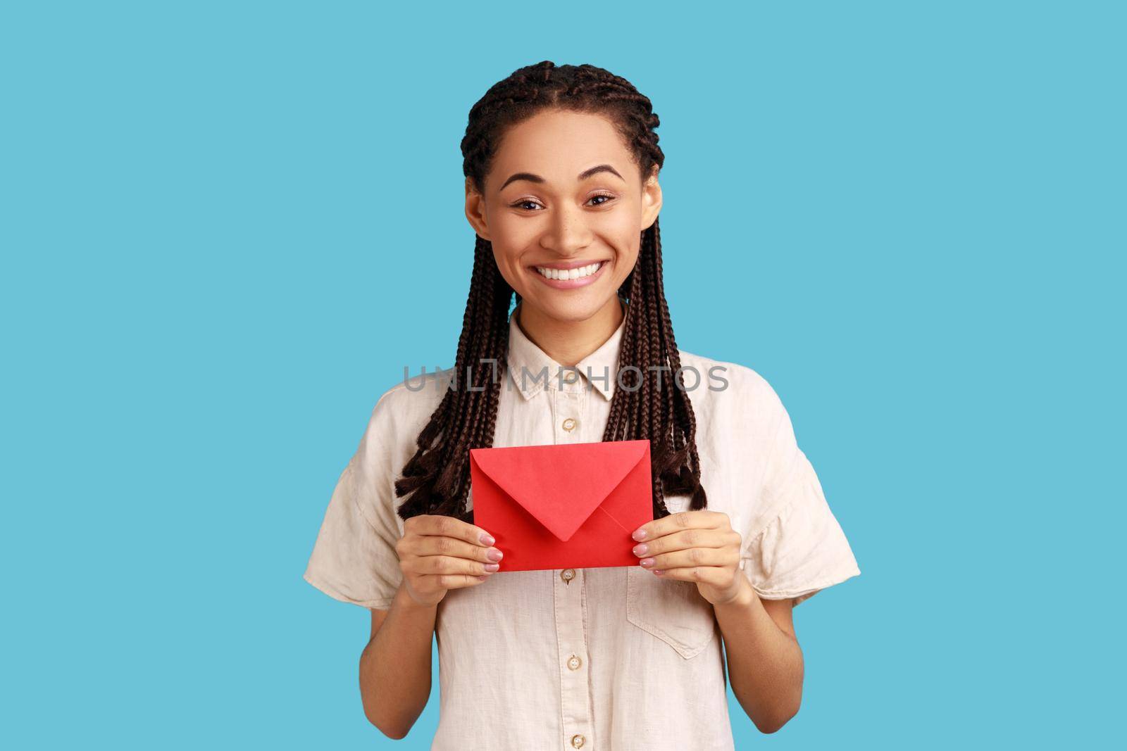 I got love letter on Valentine's day. Happy beautiful woman with black dreadlocks holding letter in red envelope or greeting card and smiling joyfully. Indoor studio shot isolated on blue background.
