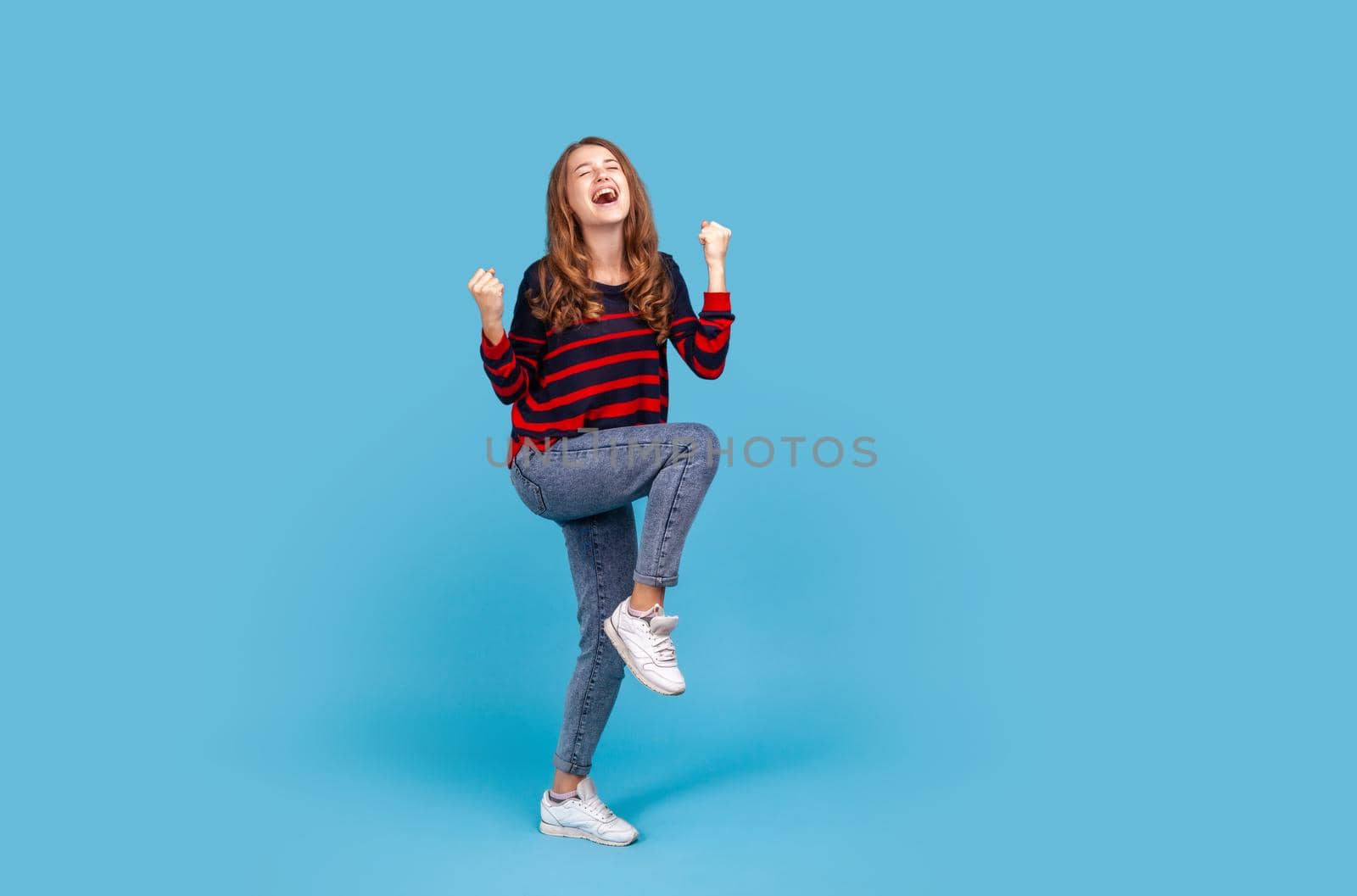 Full length positive satisfied woman wearing striped casual style sweater and jeans, raised clenched fists, being happy of winning, expressing triumph. Indoor studio shot isolated on blue background.