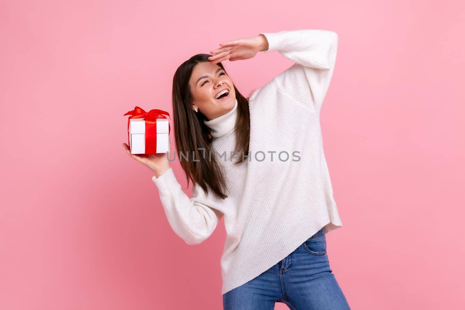 Portrait of satisfied female holding wrapped present box, looking far away with hand over forehead, wearing white casual style sweater. Indoor studio shot isolated on pink background.