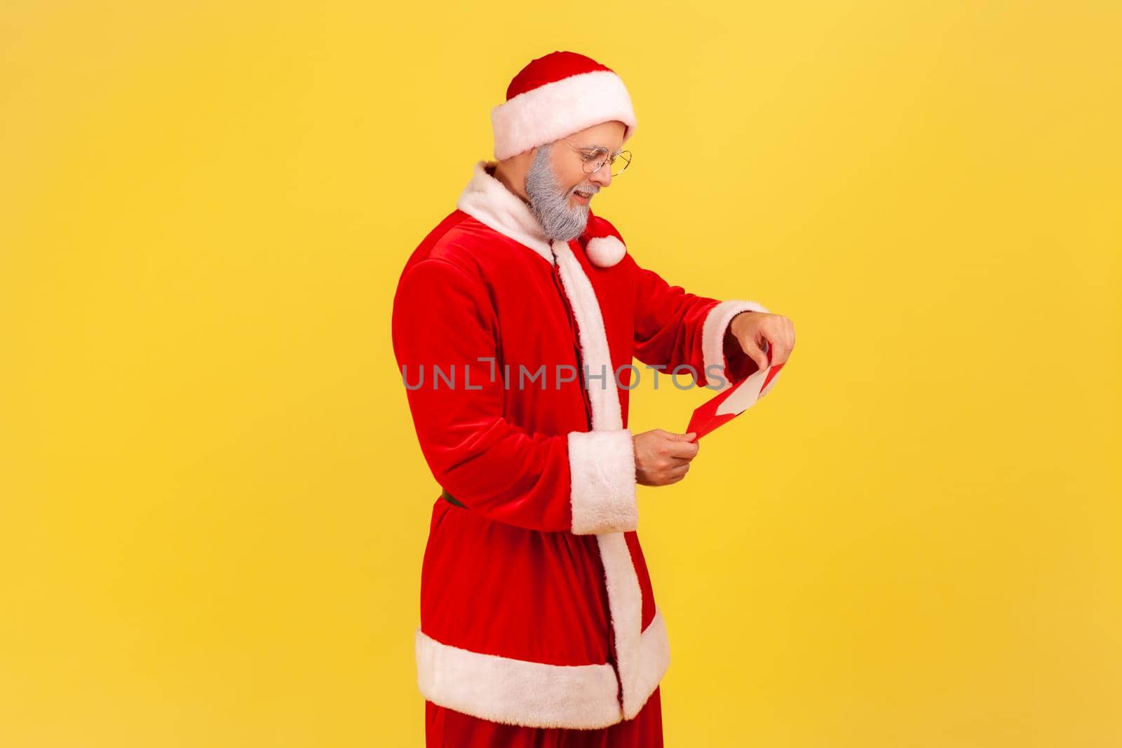 Side view of elderly man with gray beard in santa claus costume standing open red envelope with congratulations with winter holidays , reading letter. Indoor studio shot isolated on yellow background.