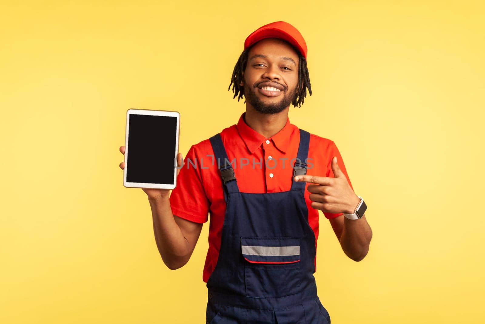 Portrait of satisfied bearded handyman wearing overalls holding tablet with empty screen for advertisement industry service, pointing at display. Indoor studio shot isolated on yellow background.