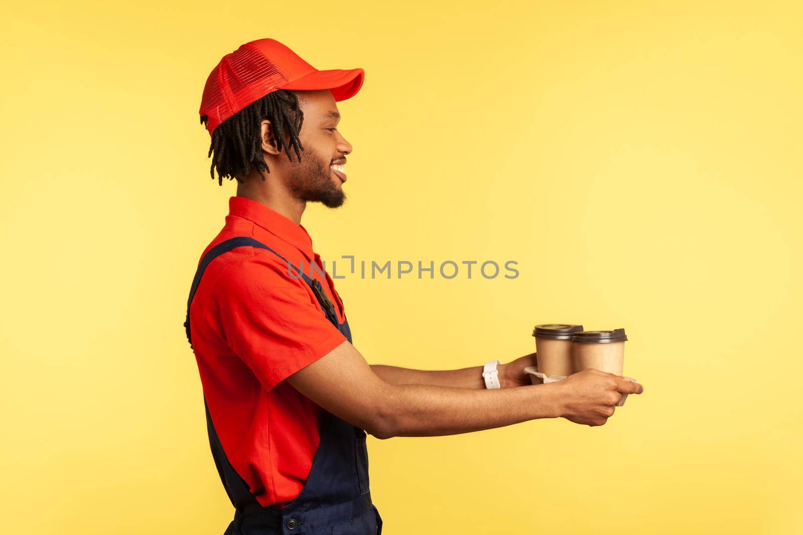 Delivery service. Side view, friendly deliveryman wearing blue overalls offering coffee, giving drinks in disposable cups and smiling. Indoor studio shot isolated on yellow background.