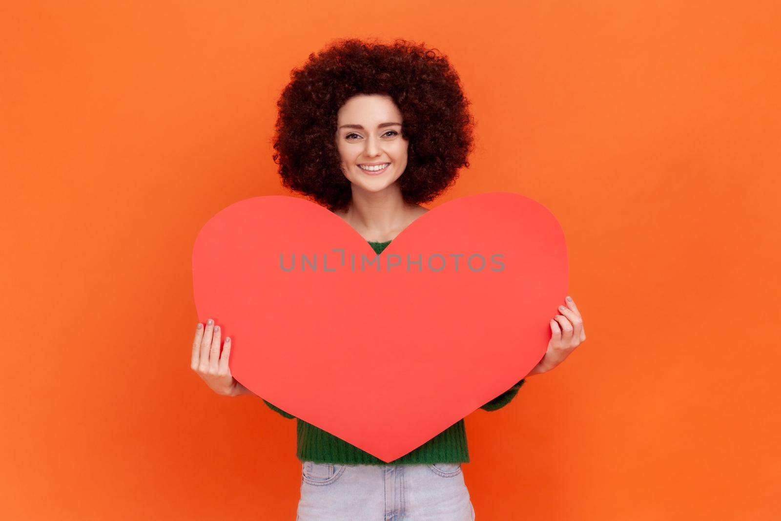 Good looking romantic woman with Afro hairstyle wearing green casual style sweater holding big red heart, expressing happiness and love. Indoor studio shot isolated on orange background.