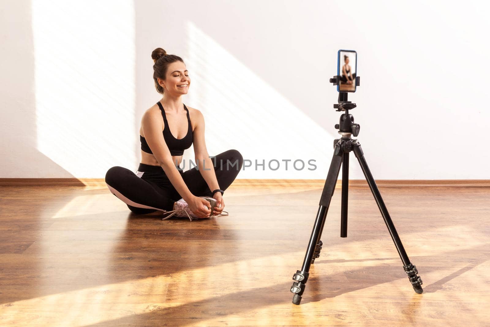 Female coach filming video of relaxing after workout, sitting on lotus pose with closed eyes, wearing black sports top and tights. Full length studio shot illuminated by sunlight from window.