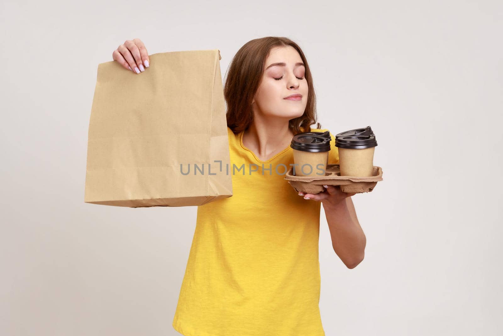 Portrait of satisfied charming teenager girl in yellow T-shirt smelling hot beverage, coffee or tea, holding paper package, tasty breakfast. Indoor studio shot isolated on gray background.