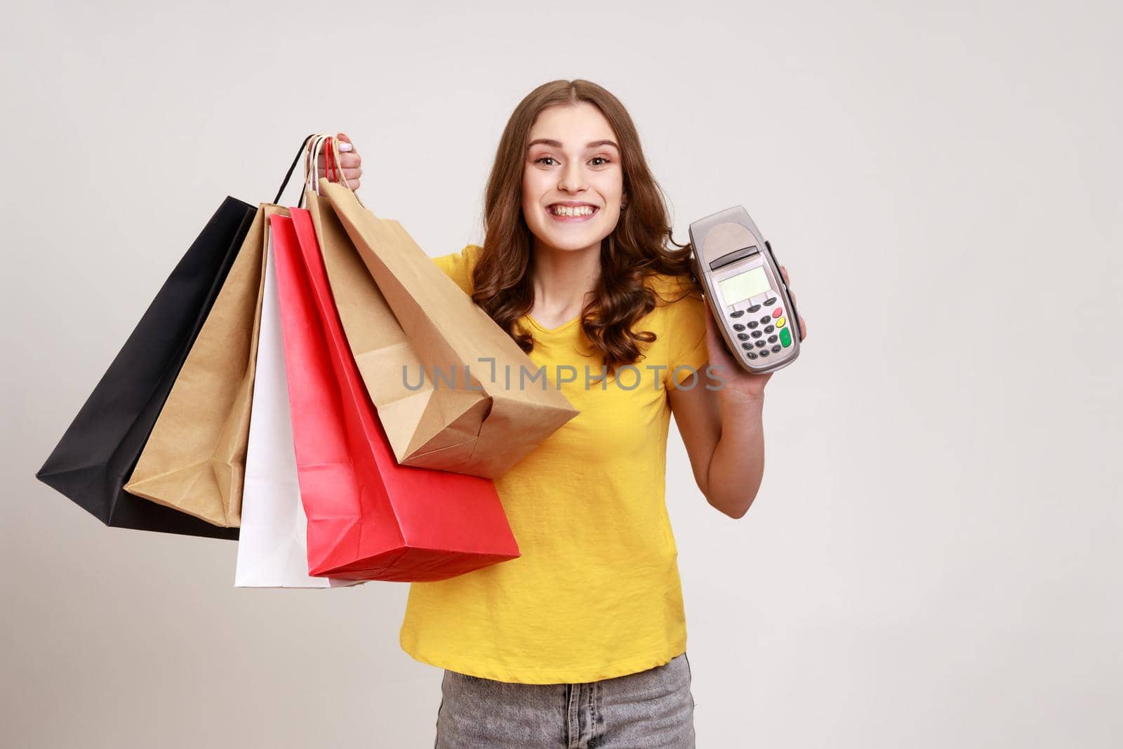 Positive smiling teenager girl with brown hair wearing yellow T-shirt holding payment terminal and paper shopping bags, easy express order and delivery. Indoor studio shot isolated on gray background.