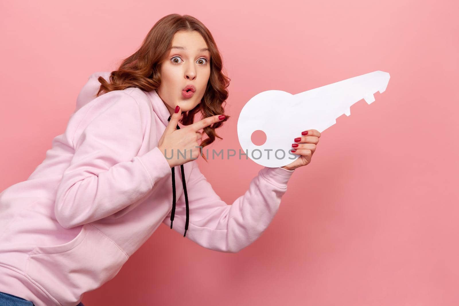 Side view portrait of curly haired teenage girl in hoodie with pout lips pointing finger on big paper key, house purchase concept. Indoor studio shot isolated on pink background