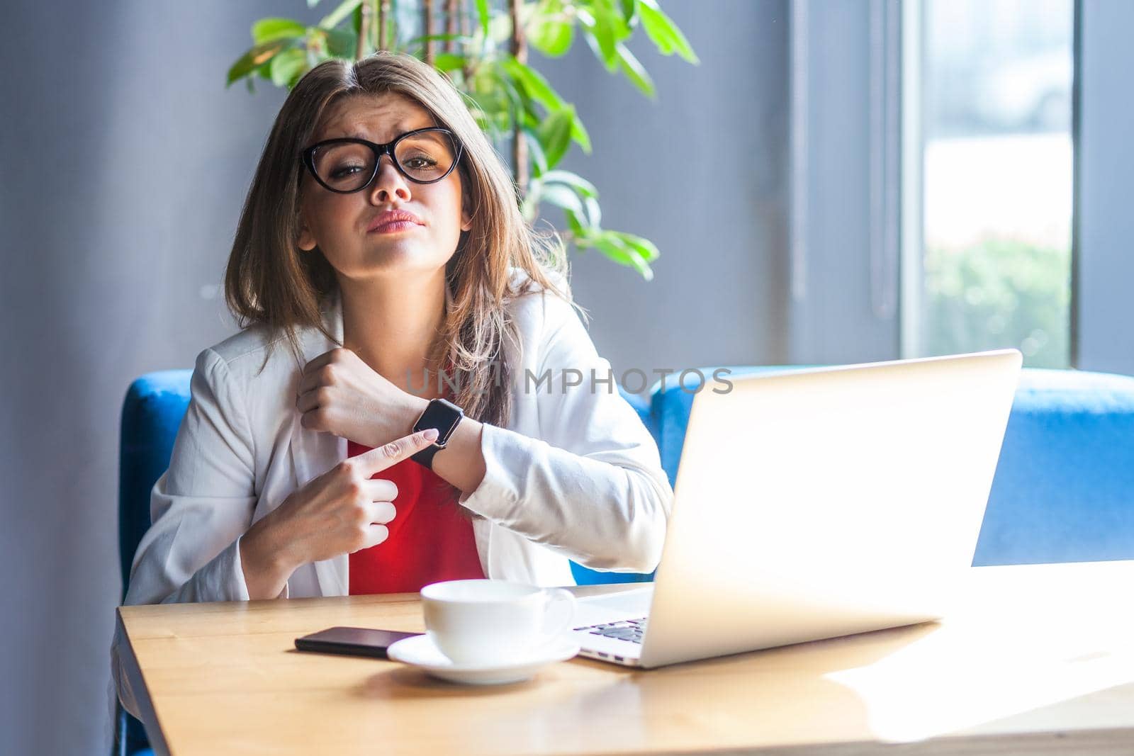 Portrait of unhappy woman in glasses pointing to smartwatch on her hand, looking gloomy disgruntled, exhausted of overtime work and need break. Indoor shot, cafe or office background.