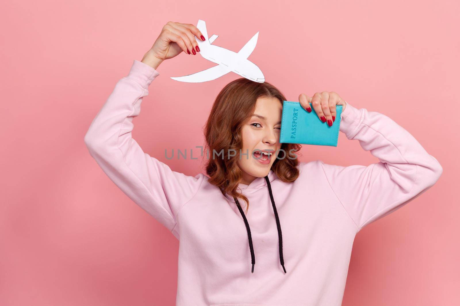 Portrait of happy curly haired teenage girl in hoodie winking, holding passport and paper airplane, looking at camera with toothy smile, travelling. Indoor studio shot isolated on pink background