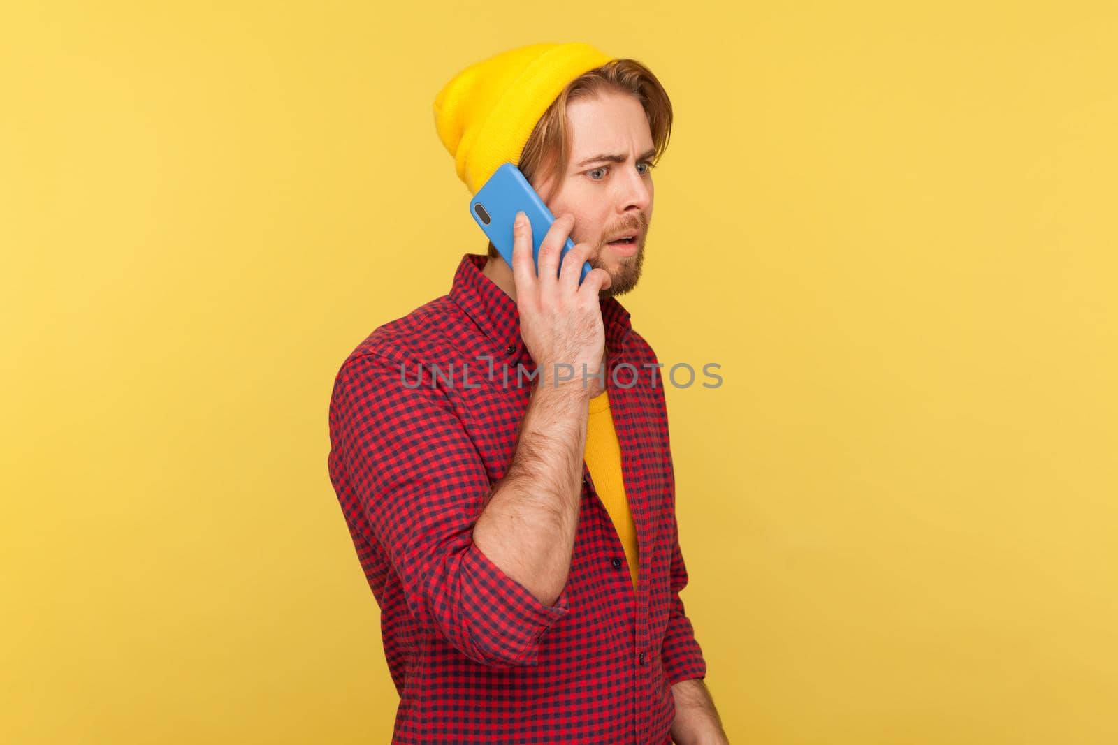 Portrait of hipster bearded guy in beanie hat and checkered shirt talking on phone, having serious mobile conversation, making important call. Indoor studio shot isolated on yellow background.