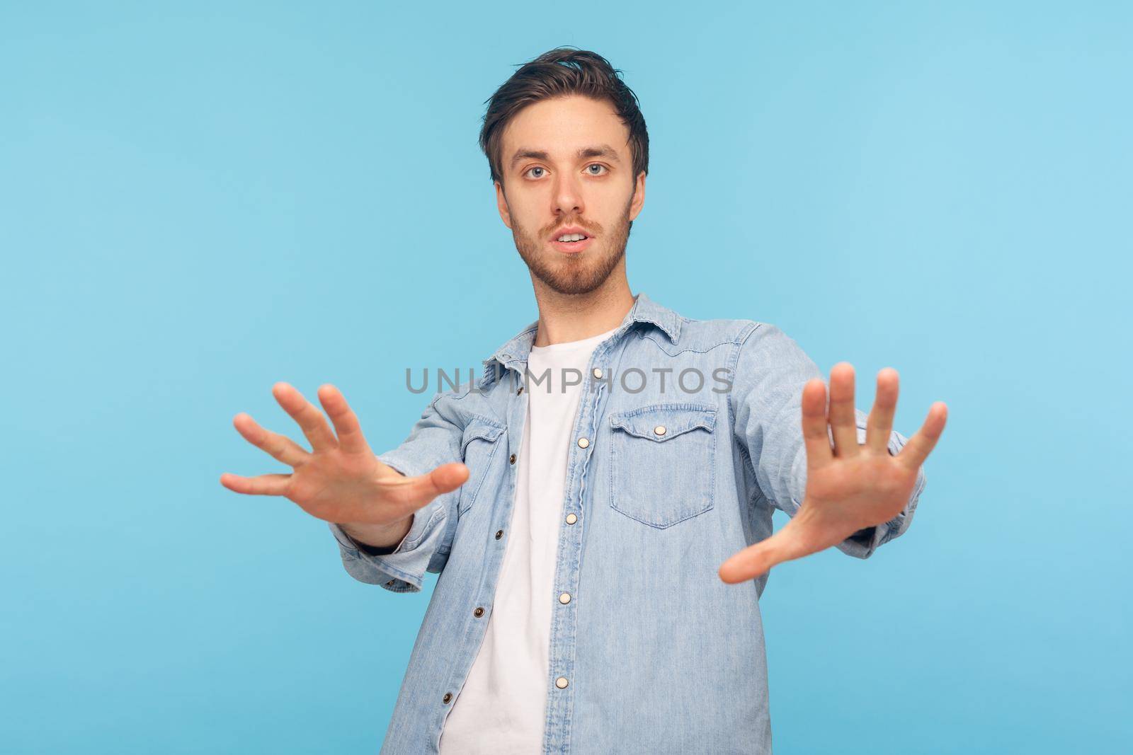 Portrait of frightened hysterical attractive man wearing denim shirt, standing, raised hands to defend from fear, horrified and shocked. Indoor studio shot isolated on blue background.
