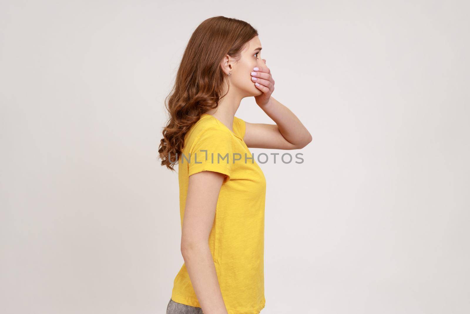 I will not say. Side view of scared bullied teen girl with brown hair in yellow t-shirt covering mouth with hand, keeping secret taboo. Indoor studio shot isolated on gray background.