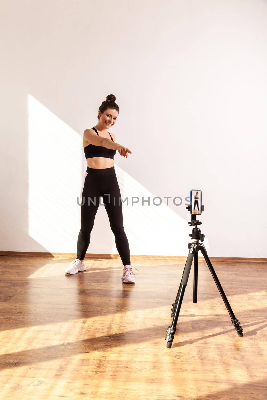 Coach conducting online sports training, pointing at phone camera on tripod, inviting you to workout by Khosro1