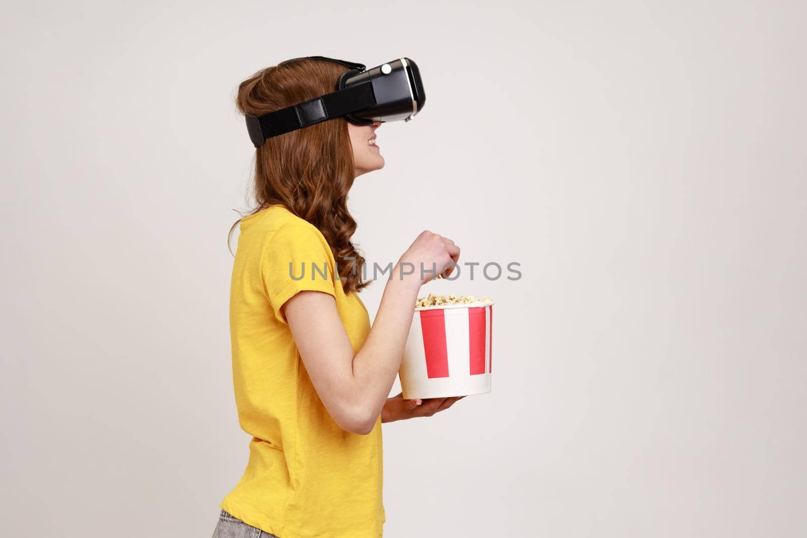 Profile portrait of young brown haired woman in VR headset watching movie with popcorn, wearing yellow casual t-shirt, eating tasty snack. Indoor studio shot isolated on gray background.