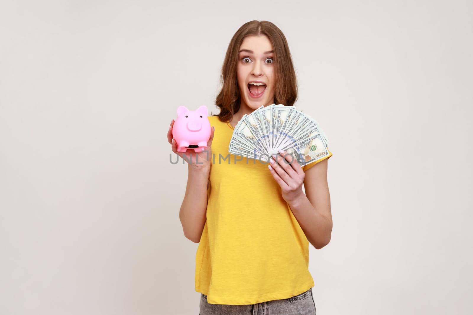 Extremely excited teen girl wearing casual style T-shirt holding piggy bank and dollar banknotes looking at camera with open mouth, deposit, cashback. Indoor studio shot isolated on gray background.