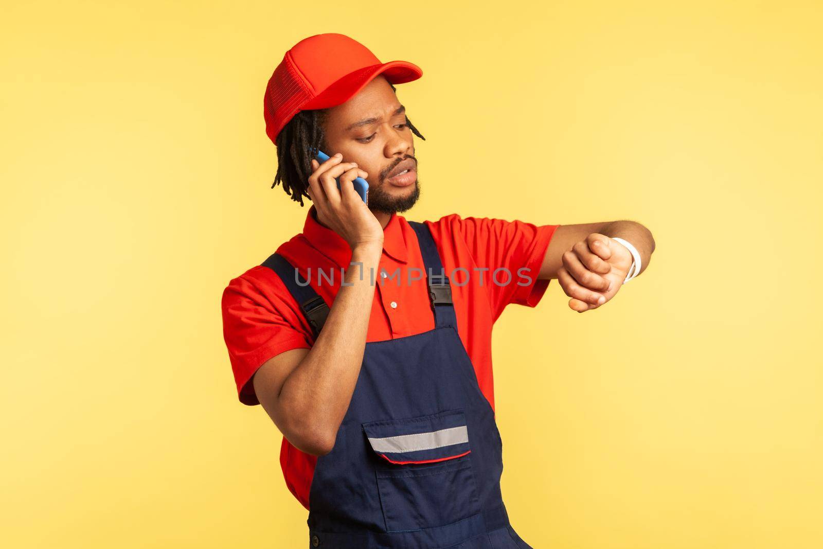 Portrait of concentrated courier wearing blue overalls talking phone with client, looking at smart watch, delivery service on time. Indoor studio shot isolated on yellow background.