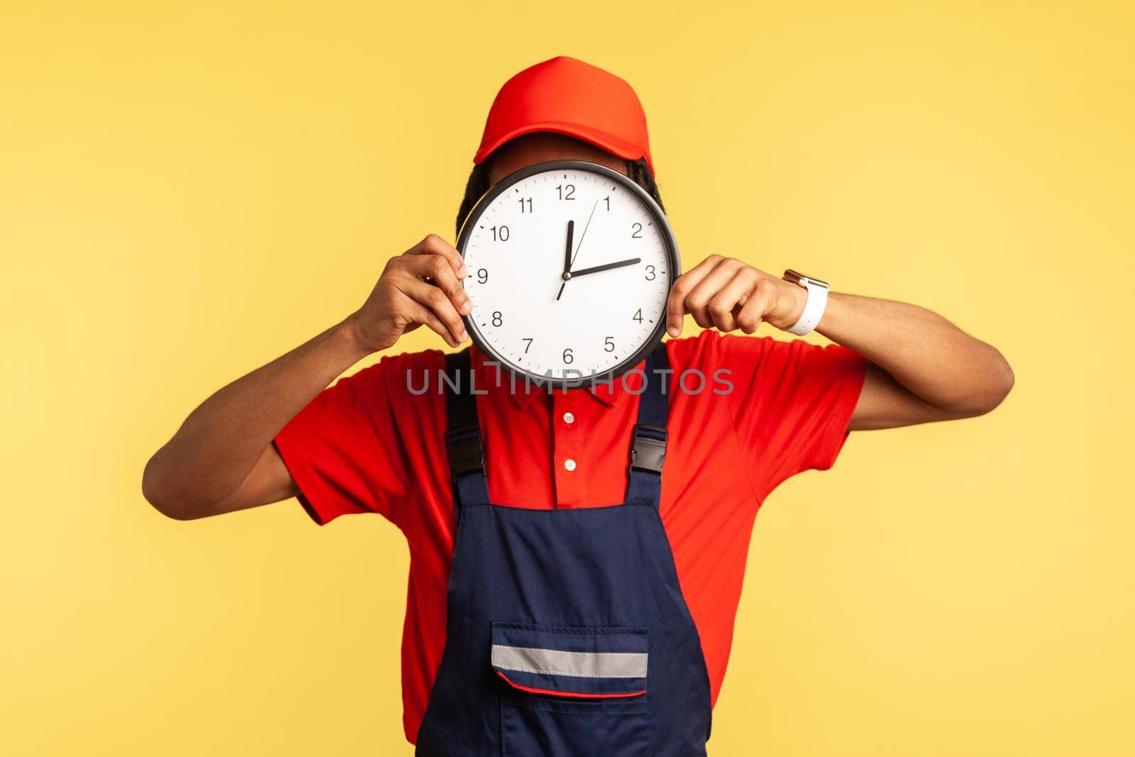 Anonymous labourer in uniform covering face with clock, advertising professional house repair services on time, punctual courier delivery. Indoor studio shot isolated on yellow background.