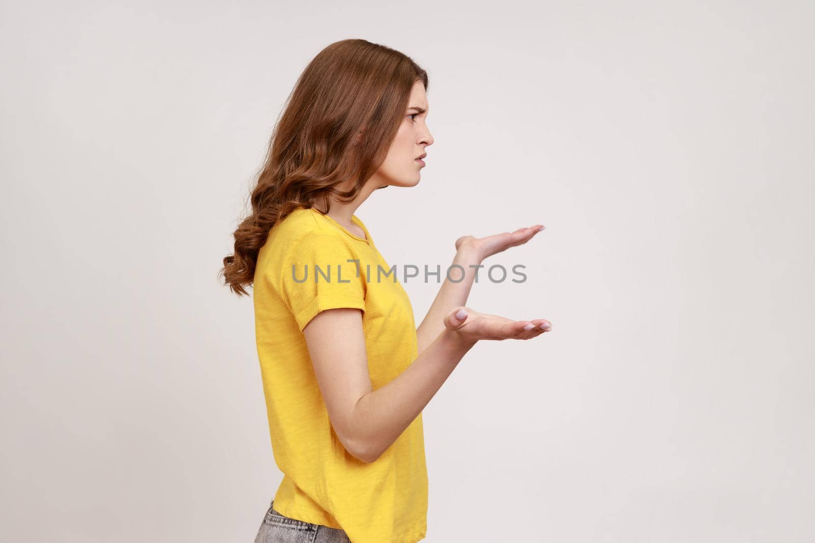 What do you want from me. Profile portrait of confused young female with brown hair in casual yellow T-shirt spreading hands, uncomfortable situation. Indoor studio shot isolated on gray background.