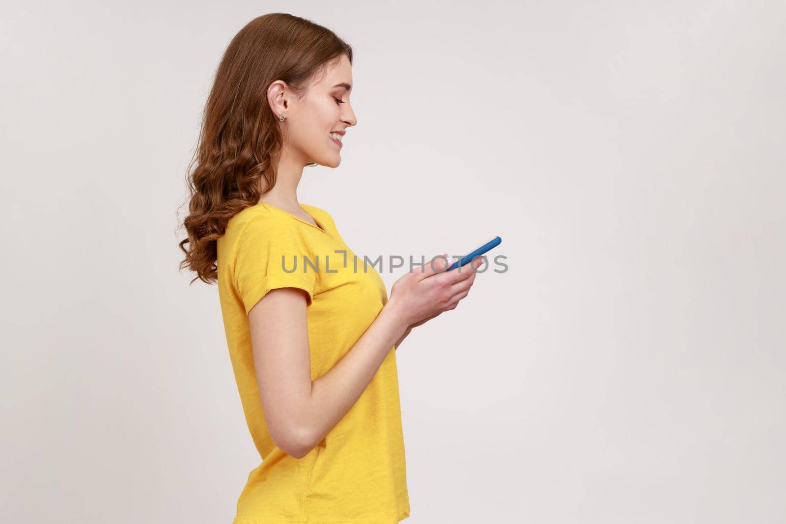 Side view portrait of smiling, happy teenager girl in yellow casual style T- shirt typing message on smartphone, looking satisfied pleased with app. Indoor studio shot isolated on gray background.