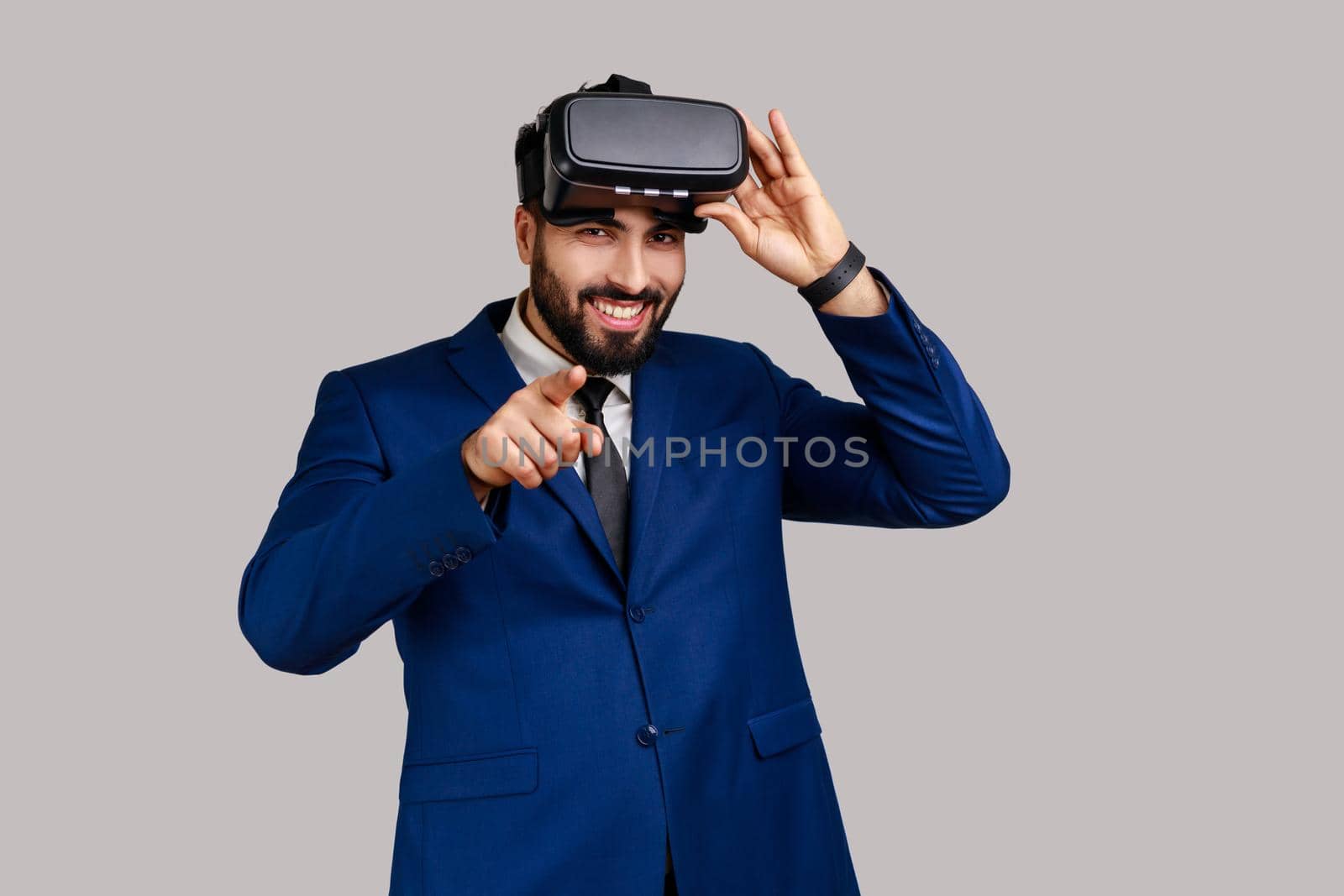 Smiling man with virtual reality glasses expressing happiness, being ready for playing game simulator, pointing to camera, choosing you as opponent. Indoor studio shot isolated on gray background.