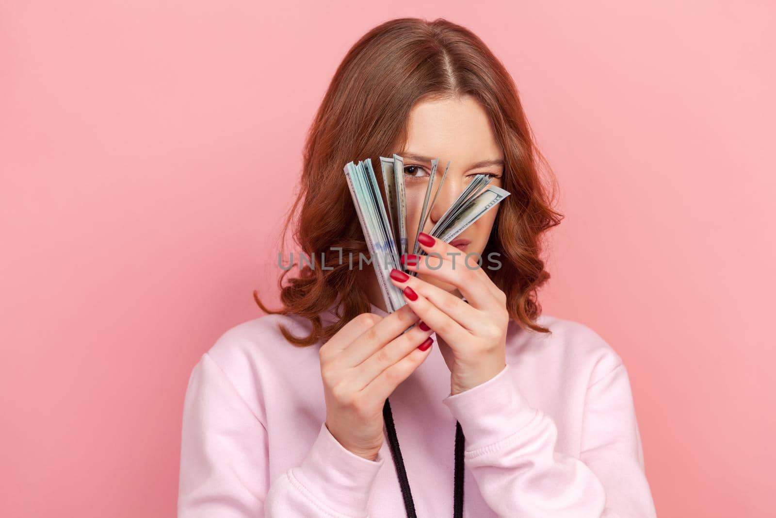 Portrait of curious curly haired teenage girl in hoodie peeping through bunch of dollar banknotes, passive income by Khosro1