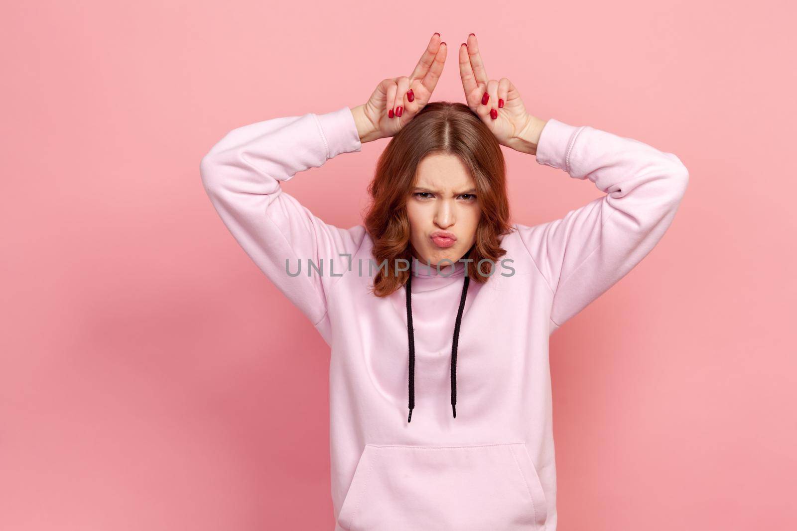 Portrait of aggressive teen girl in hoodie standing with bull horns, antler head gesture, threatening to attack, conflict or defence concept by Khosro1