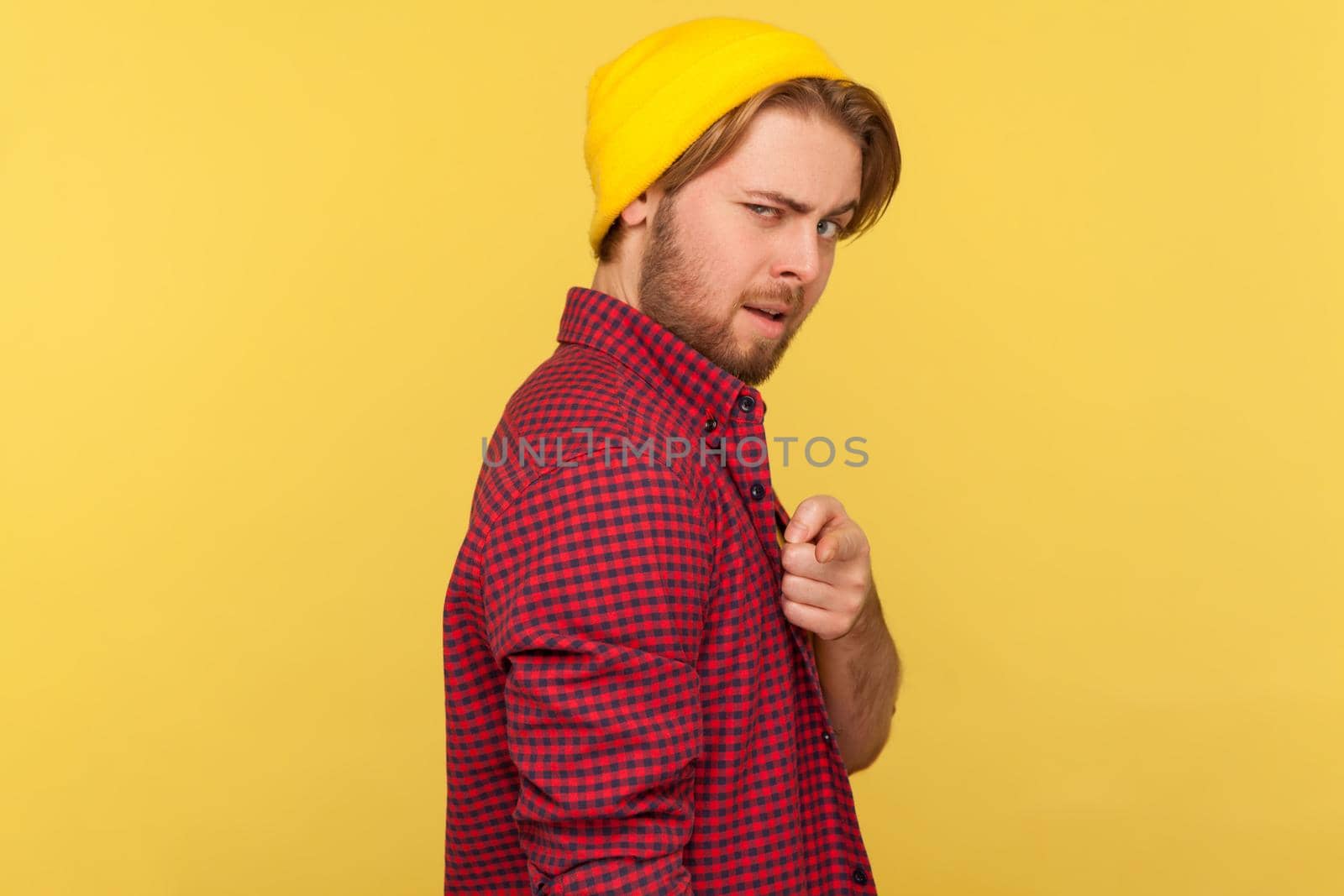 Portrait of guy in beanie hat and checkered shirt pointing finger at camera with flirting expression, choosing you, saying you are what we need. Indoor studio shot isolated on yellow background.